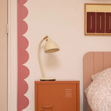 Roze kinderkamermuur met een golvende behangrand van Bibelotte naast een bed en terracotta nachtkastje